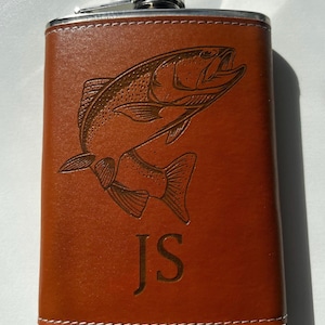 Puede incluir: Una petaca de cuero marrón con tapa plateada y un grabado detallado de un pez. La petaca tiene las iniciales "JS" grabadas en la parte delantera. La petaca es rectangular y tiene costuras blancas en los bordes.