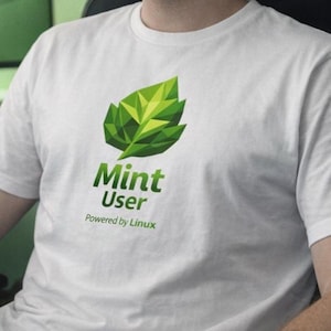 May include: White t-shirt featuring a green geometric leaf design and the text "Mint User Powered by Linux". The shirt is made of a soft, light-colored fabric and is suitable for casual wear.