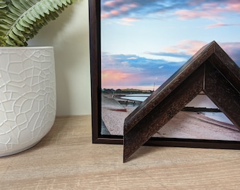 May include: A dark brown wood picture frame with a beveled edge. The frame is holding a canvas print of a beach scene with a sunset sky.