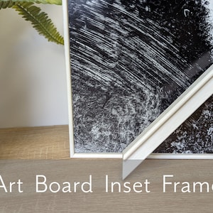 May include: White art board inset frame with a black and white abstract print. The frame is on a light wood surface.