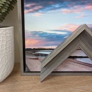May include: A framed canvas print of a seascape with a pink and blue sunset over a beach. The frame is a light grey wood with a bevelled edge.