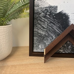 May include: A dark brown wood picture frame with a black and white abstract painting inside. The frame is on a light brown wooden surface.