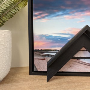 May include: A black wood frame with a bevelled edge surrounds a canvas print of a beach scene with a pink and blue sunset sky.