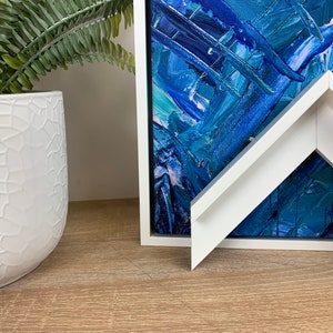 May include: An abstract painting in shades of blue and white, presented in a white frame. A white, textured ceramic pot with a green plant is positioned nearby, creating a modern aesthetic.