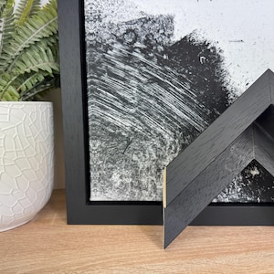 May include: A black picture frame with a black and white abstract painting inside. The frame has a wide, black mat and a simple, modern design.