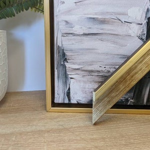May include: A gold-toned picture frame with a woodgrain finish. The frame is angled to show the detail of the frame's edge. The frame is on a wooden surface.