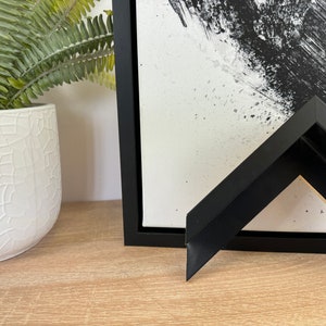 May include: Abstract black and white artwork in a black frame. The painting features bold brushstrokes and splatters. A separate black frame corner is displayed in the foreground, showcasing the frame's design. A white ceramic pot with a green plant is on the left.
