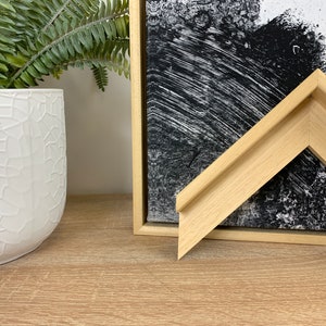 May include: A close-up of a natural wood picture frame with a bevelled edge. The frame is light brown in colour and has a simple, modern design.