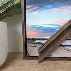 May include: A framed canvas print of a beach scene with a cloudy sky at sunset. The frame is made of dark wood with a visible wood grain.
