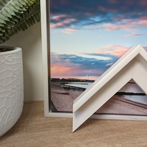 May include: A white picture frame with a bevelled edge and a canvas print of a beach scene with a cloudy sky. The frame is sitting on a wooden surface.