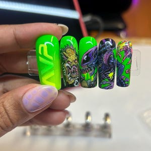 May include: A set of five long, neon green artificial nails with various designs. One nail features the letters "VR" in a gradient, while the others display monster-themed illustrations in black, purple, and green.