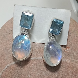 Könnte beinhalten: Ein Paar silberne Ohrhänger mit einem hellblauen Edelstein und einem weißen Mondstein. Die Ohrringe sind auf einer weißen Karte und einem braunen Stein abgebildet.