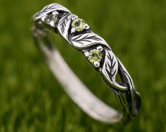 May include: A silver ring with a leaf and vine design. The ring features three small, round, light green gemstones. The band is detailed with leaves and vines, creating a nature-inspired aesthetic. The ring is set against a green background.