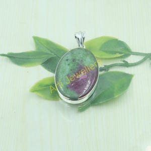 Könnte beinhalten: Ovaler Anhänger mit einem grünen und roten Edelstein, der in einen silberfarbenen Metallrahmen gefasst ist. Der Anhänger hat eine Öse für eine Kette. Der Text "Arti Jeweller" ist sichtbar.