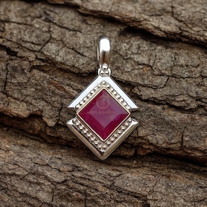 以下が含まれることがあります： 正方形の濃い赤色の宝石がダイヤモンド型のフレームにセットされたシルバーペンダント。ペンダントの上部にはチェーン用の小さなループがあり、木目調の茶色の木材の表面に置かれています。「Arti Jewellers」の文字が見えます。