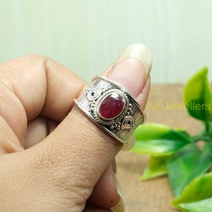 Może przedstawiać: Srebrny pierścionek z czerwonym kamieniem szlachetnym osadzonym w ozdobnej srebrnej oprawie. Pierścionek ma fakturowany pasek z misternymi detalami.