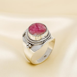 May include: A silver ring with a large, oval pink gemstone. The gemstone is set in a raised bezel, encircled by a twisted rope design. The ring has a wide band with decorative details, including small silver beads.