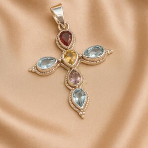 May include: A silver pendant in the shape of a cross, adorned with teardrop-shaped gemstones in various colors: red, yellow, light blue, and purple. The pendant has a textured border and a loop for a chain.