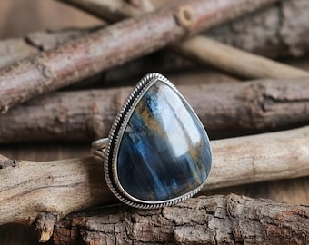 May include: A teardrop-shaped ring featuring a large, polished blue gemstone with gold and white veining. The ring has a silver-coloured, textured band. The gemstone is approximately 3.8 cm long, set against wooden branches.