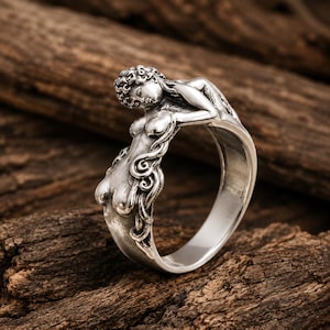 May include: A silver ring featuring a sculpted figure of a reclining person with detailed hair and body contours. The metal has a polished finish with darkened recessed areas to highlight the intricate relief design of the human form.