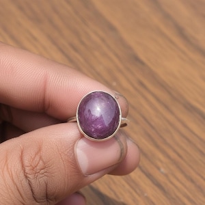 May include: A silver ring featuring a large, oval-shaped, deep purple gemstone. The gemstone has a polished, reflective surface. The ring is held by a hand against a blurred wooden background.