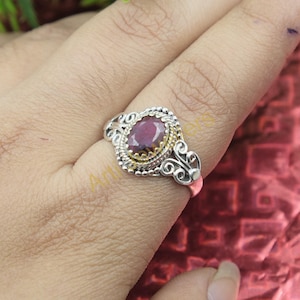 May include: A silver ring with an oval, deep red gemstone. The gemstone is set in a gold-colored bezel, surrounded by a silver filigree design. The ring is on a finger, with a red background.