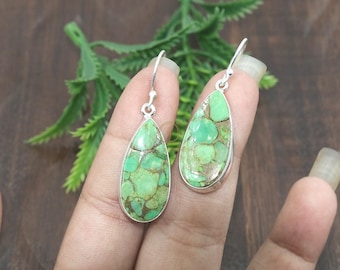 May include: A pair of teardrop-shaped earrings with a silver setting. The earrings have a green stone with brown and gold veining. The earrings are shown against a dark wooden background with green foliage.
