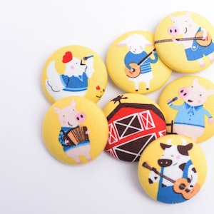 May include: Six colorful fabric buttons with farm animal designs. Each button features a different animal playing a musical instrument, including a pig playing an accordion, a cow playing a guitar, a pig playing a banjo, a sheep playing a banjo, a pig singing into a microphone, and a rooster holding a microphone. The buttons are yellow with red, blue, and brown accents.