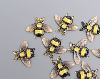 May include: A collection of ten decorative bee charms. Each bee features a black and yellow body, bronze-colored wings, and a detailed design. The charms are arranged on a gray surface.