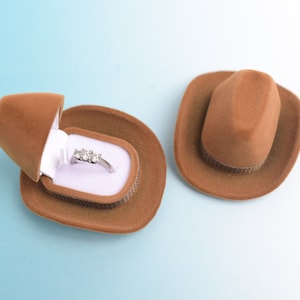 May include: A brown cowboy hat-shaped ring box, open to reveal a silver ring with three stones. Another closed hat is next to it. The ring box is on a light blue background.