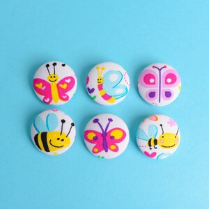 May include: Six round fabric buttons with colorful butterfly and bee designs. The buttons feature white backgrounds with pink, yellow, blue, and purple illustrations. The buttons are arranged on a light blue background.