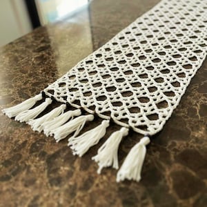 May include: A white crocheted table runner with a diamond pattern and fringe tassels. The runner is on a brown marble surface.