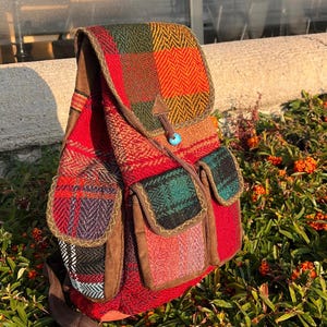 Può includere: Uno zaino rosso e a quadri con cinghie e tasche marroni. Lo zaino ha una chiusura con patta e un bottone turchese. La borsa è appoggiata su un letto di piante verdi con bacche arancioni.