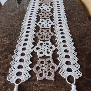 May include: A cream and beige crochet table runner with a floral pattern. The runner has a repeating design of open circles and flower shapes. Tassels are attached at each end, providing a decorative element to this handmade textile.