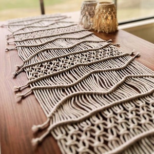 Puede incluir: Un camino de mesa de macramé beige con un patrón tejido. El camino de mesa está sobre una mesa de madera con dos pequeños candelabros.