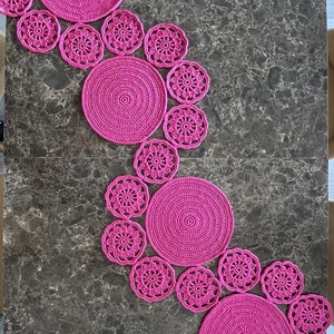 Puede incluir: Un juego de manteles individuales y posavasos de crochet de color rosa. Los manteles individuales son grandes y redondos, mientras que los posavasos son más pequeños y tienen un diseño floral.