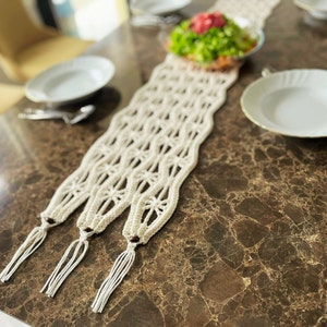 Könnte beinhalten: Cremefarbener Makramee-Tischläufer mit geometrischem Muster und Quasten. Auf einem braunen Tisch mit Marmoroptik, mit weißen Tellern und Besteck. Im Hintergrund eine Schüssel mit Salat. Der Tischläufer ist ca. 150 cm lang.