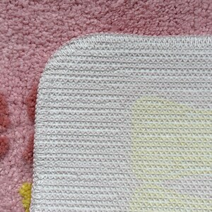May include: Close-up of a pink rug with a floral design. The rug has a soft, textured surface and a white, non-slip backing with a geometric pattern. The edges are neatly stitched with white thread.