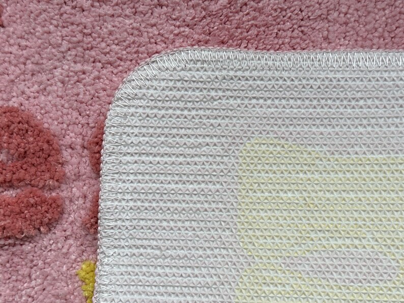 May include: Close-up of a pink rug with a floral design. The rug has a soft, textured surface and a white, non-slip backing with a geometric pattern. The edges are neatly stitched with white thread.