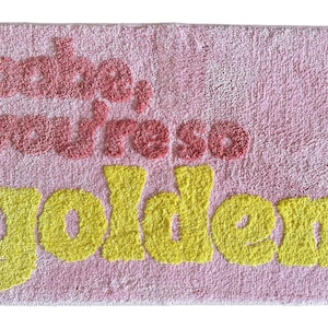 May include: Pink rectangular bath mat with the words "babe, you're so golden" in red and yellow. The mat has a textured, plush appearance, and the text is in a playful, bubbly font.