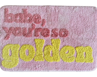 Puede incluir: Alfombra de baño rectangular rosa con las palabras "babe, you're so golden" en rojo y amarillo. La alfombra tiene un aspecto texturizado y afelpado, y el texto está en una fuente juguetona y burbujeante.