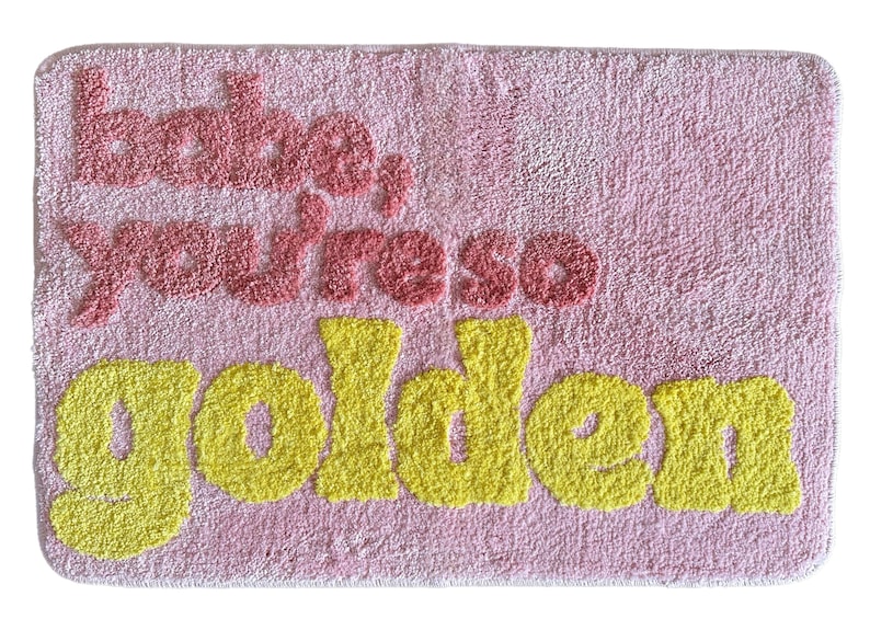 May include: Pink rectangular bath mat with the words "babe, you're so golden" in red and yellow. The mat has a textured, plush appearance, and the text is in a playful, bubbly font.
