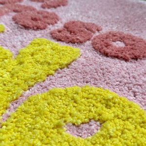 May include: Close-up of a pink rug with yellow and red floral designs. The rug has a textured, tufted appearance. The yellow flowers are in the foreground, with red flowers and text in the background.