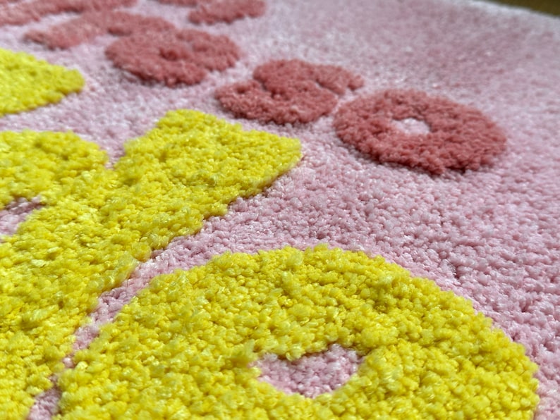 May include: Close-up of a pink rug with yellow and red floral designs. The rug has a textured, tufted appearance. The yellow flowers are in the foreground, with red flowers and text in the background.