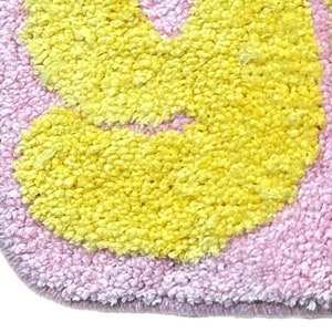 May include: Close-up of a soft, plush rug with a floral design. The rug features a light pink background with yellow flower shapes. The edges are a darker shade of pink, creating a border.
