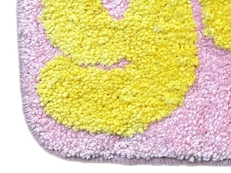 May include: Close-up of a soft, plush rug with a floral design. The rug features a light pink background with yellow flower shapes. The edges are a darker shade of pink, creating a border.