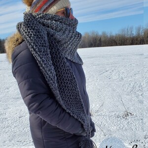 May include: A gray, chunky-knit scarf with a matching tassel, worn with a dark blue winter coat and a striped knit hat. The scarf is wrapped around the neck and face. The background is a snow-covered field under a blue sky.