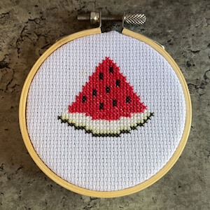 May include: A white cross-stitch embroidery hoop with a red watermelon design. The watermelon is stitched with red, white, and green thread.