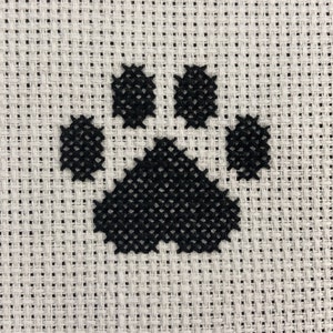 May include: A black paw print cross-stitched on white Aida cloth.