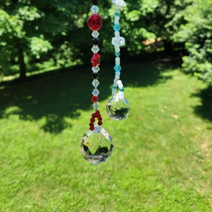 May include: Two crystal suncatchers hanging from a string of beads. The suncatchers are clear and have a faceted design. The beads are red, black, white, and blue.